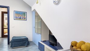 Une pièce avec un lit recouvert d’une couverture bleue, un bureau avec une télévision, une horloge fixée au mur et un panier de citrons posé sur une table.