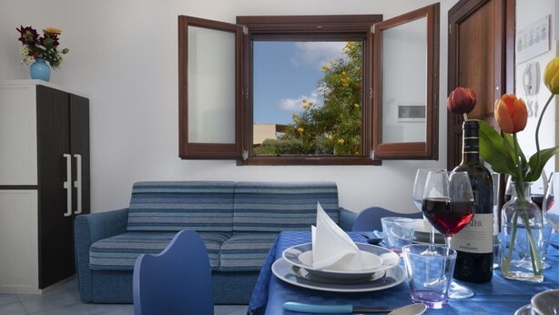 Un coin repas avec une table dressée pour deux personnes, un canapé, un meuble et une fenêtre donnant sur un paysage verdoyant.