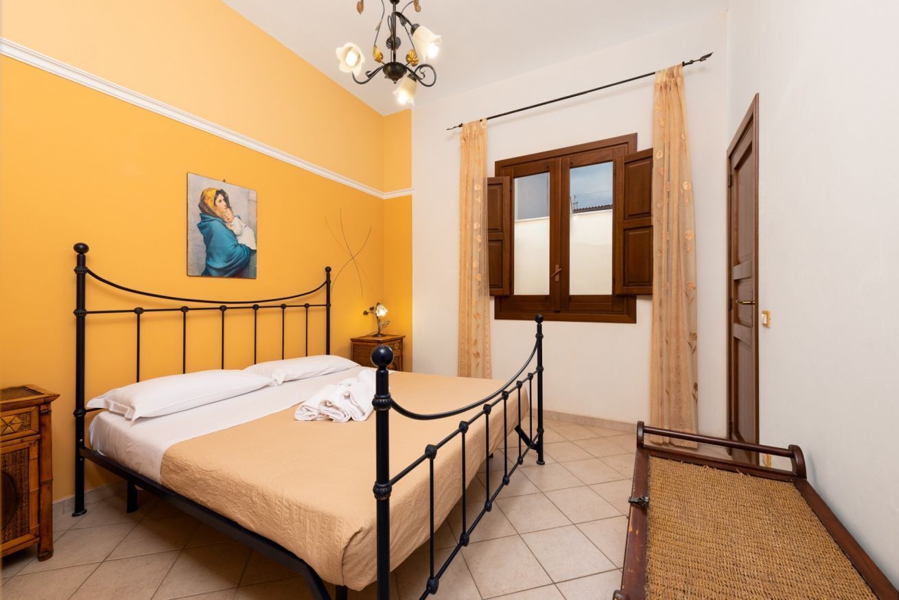 Un dormitorio con cama, mesita de noche, un cuadro, ventana con cortinas y puerta.
