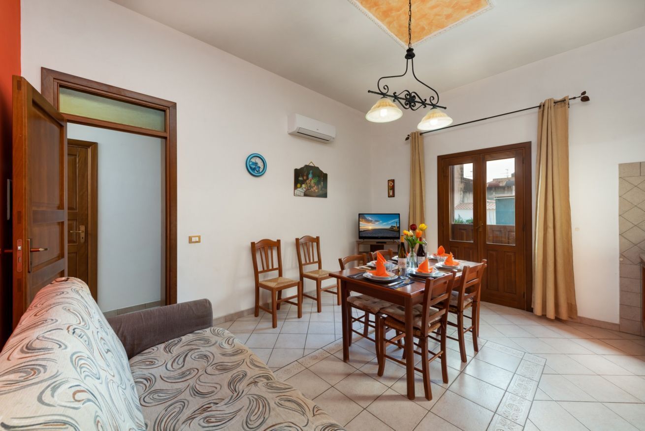 Una sala de estar con sofá, mesa de comedor, sillas y televisor. Se observa una puerta, una ventana y una lámpara del techo.