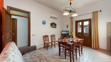 Una sala de estar con sofá, mesa de comedor, sillas y televisor. Se observa una puerta, una ventana y una lámpara del techo.