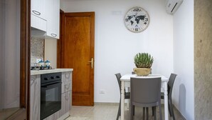 A compact kitchen with a stove, oven, and a dining table with chairs. A wooden door and a wall clock are also visible.