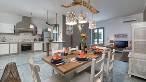 A modern kitchen with white cabinets, a wooden dining table set for four, and a large window. - I Tre Golfi Villa Iride (San Vito Lo Capo)