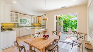 Une cuisine moderne avec des meubles blancs, une table à manger en bois avec des chaises, et une vue sur un jardin verdoyant par la baie vitrée coulissante.