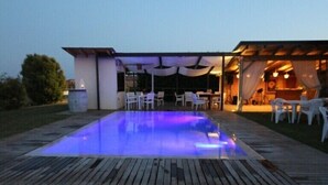 An outdoor swimming pool with a wooden deck, surrounded by white chairs and tables, and a covered area with a stage and lighting. - Osteria e Locanda del Viaggiatore (Russi)