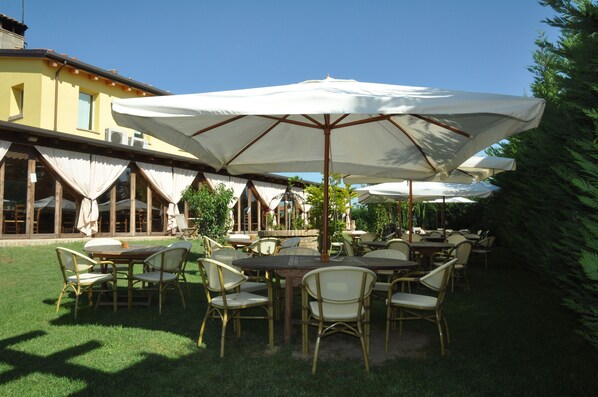 An outdoor patio with tables and chairs, covered by large umbrellas, and surrounded by greenery.