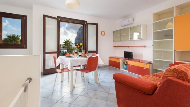 Ein modernes Wohnzimmer mit einem roten Sofa, ein Essbereich mit einem weißen Tisch und orangefarbenen Stühlen sowie eine Küche mit Fernseher und Schränken.