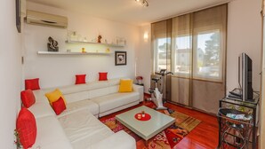 A living room with a white sofa, red cushions, a coffee table, a TV, and a window with blinds. - Apartments Zenta (Split)