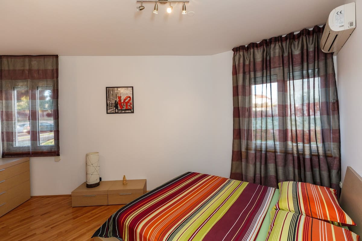 A bedroom with a bed, a bedside table, a dresser, a window with curtains, an air conditioner, and a framed picture on the wall.