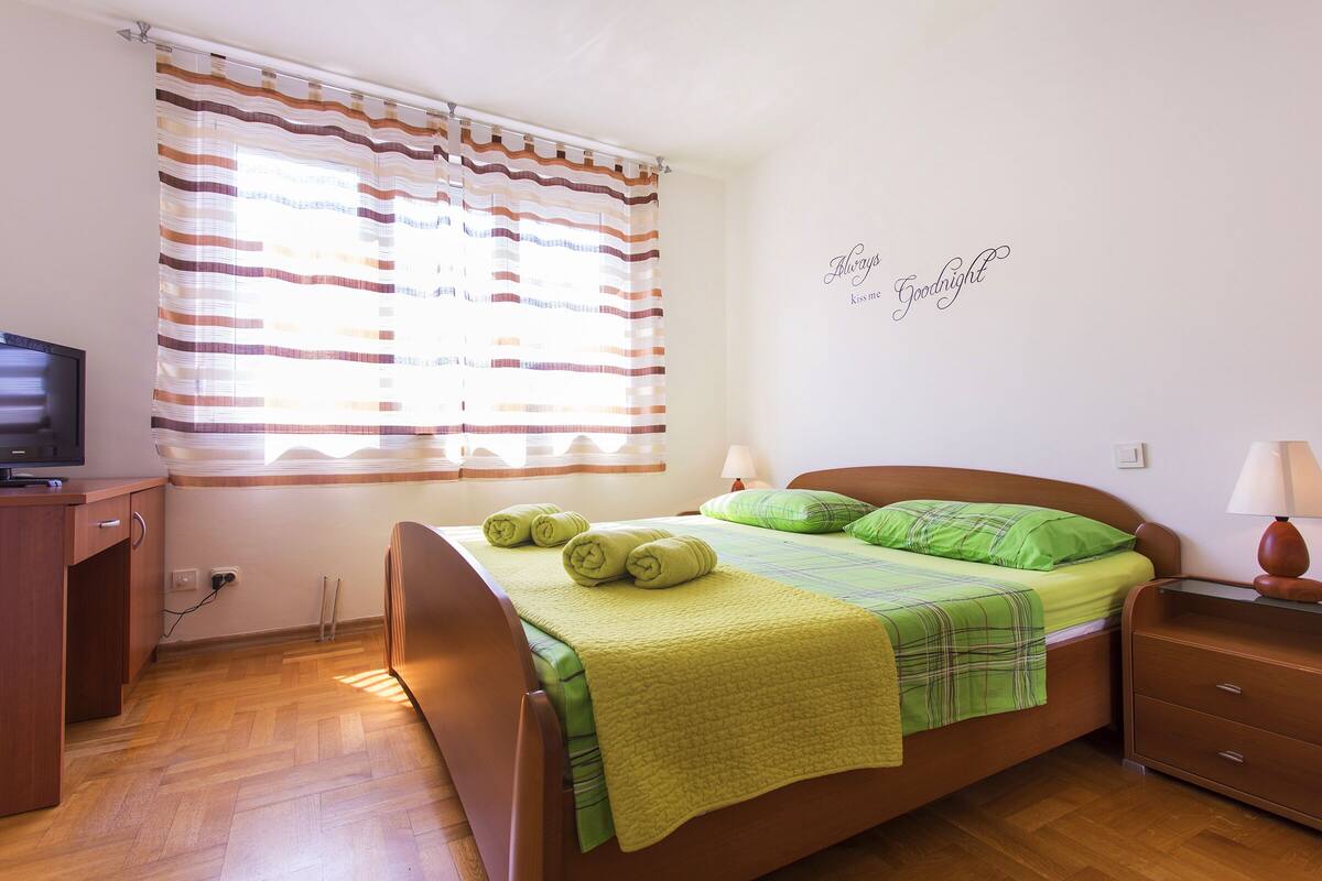 A neatly made bed with a green blanket and pillows, a bedside table with a lamp, and a television on a wooden desk.