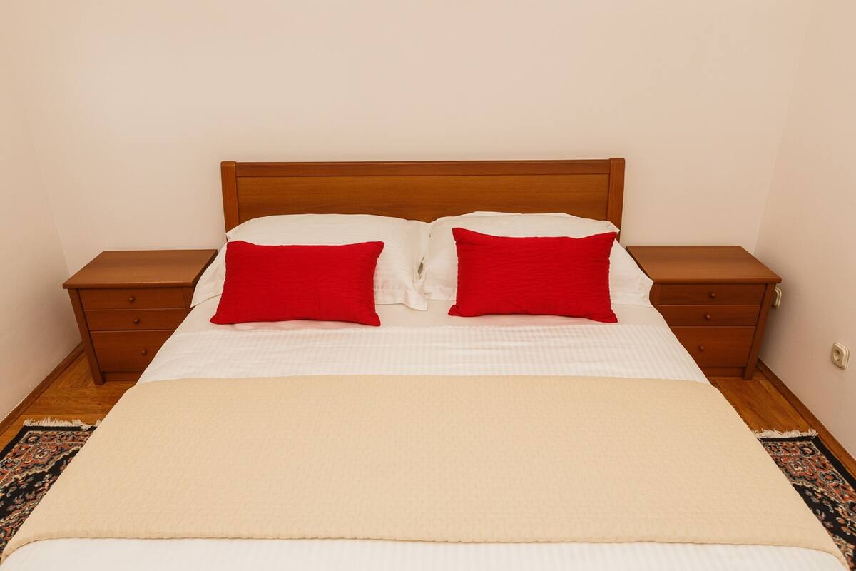 A neatly made bed with a wooden headboard, two red pillows, and a beige blanket.