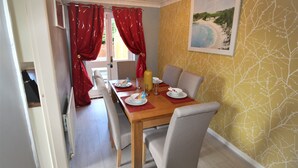 A dining area with a wooden table set for four, red curtains, and a painting of a beach on the wall. - DO NOT USE (Doncaster)
