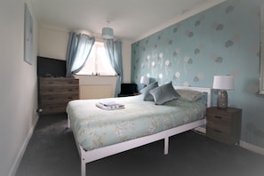 Quarto com cama, penteadeira, mesa de cabeceira, luminária e televisão.