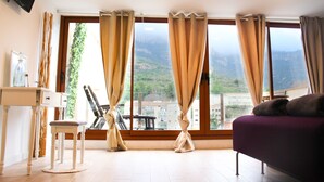 Une pièce avec une grande fenêtre, un canapé, un fauteuil, une petite table et un téléviseur fixé au mur.