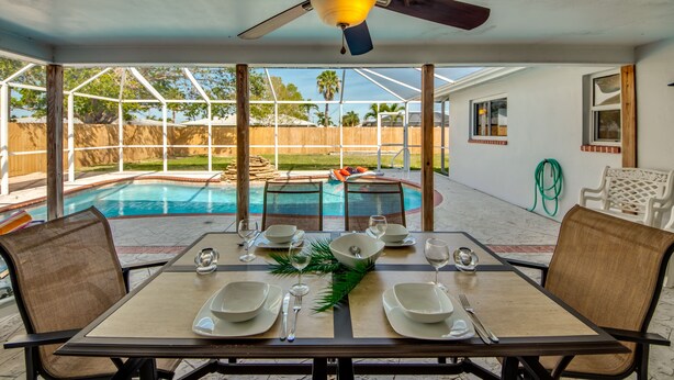 A dining area with a table set for four, a ceiling fan, and a view of a backyard with a pool. - 5 O'clock Somewhere - Private Elec. Heated Pool - WIFI - Streaming TV - BBQ (Cape Coral)