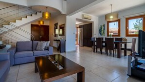 A spacious living area with a sofa, coffee table, and a dining area with chairs and a TV. A staircase with a unique design leads to another level. - Villa 148 Lofou - Four Bedroom Villa, Sleeps 8 (Kouklia)