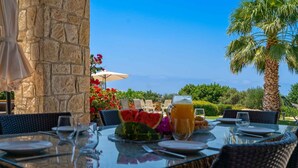 An outdoor dining area with stone walls, wicker furniture, and a view of the sea. - Villa 148 Lofou - Four Bedroom Villa, Sleeps 8 (Kouklia)