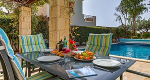 Mesa de vidrio al aire libre con cojines a rayas, una piscina y un muro de piedra.