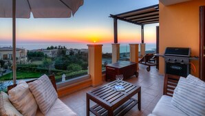 A balcony with a view of the sunset, a grill, and outdoor furniture.