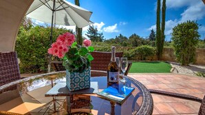 A glass table with a bottle of wine, a glass, and a book. There is a wicker chair and a white umbrella. - Apartment APD02 - Artemis (Kouklia)