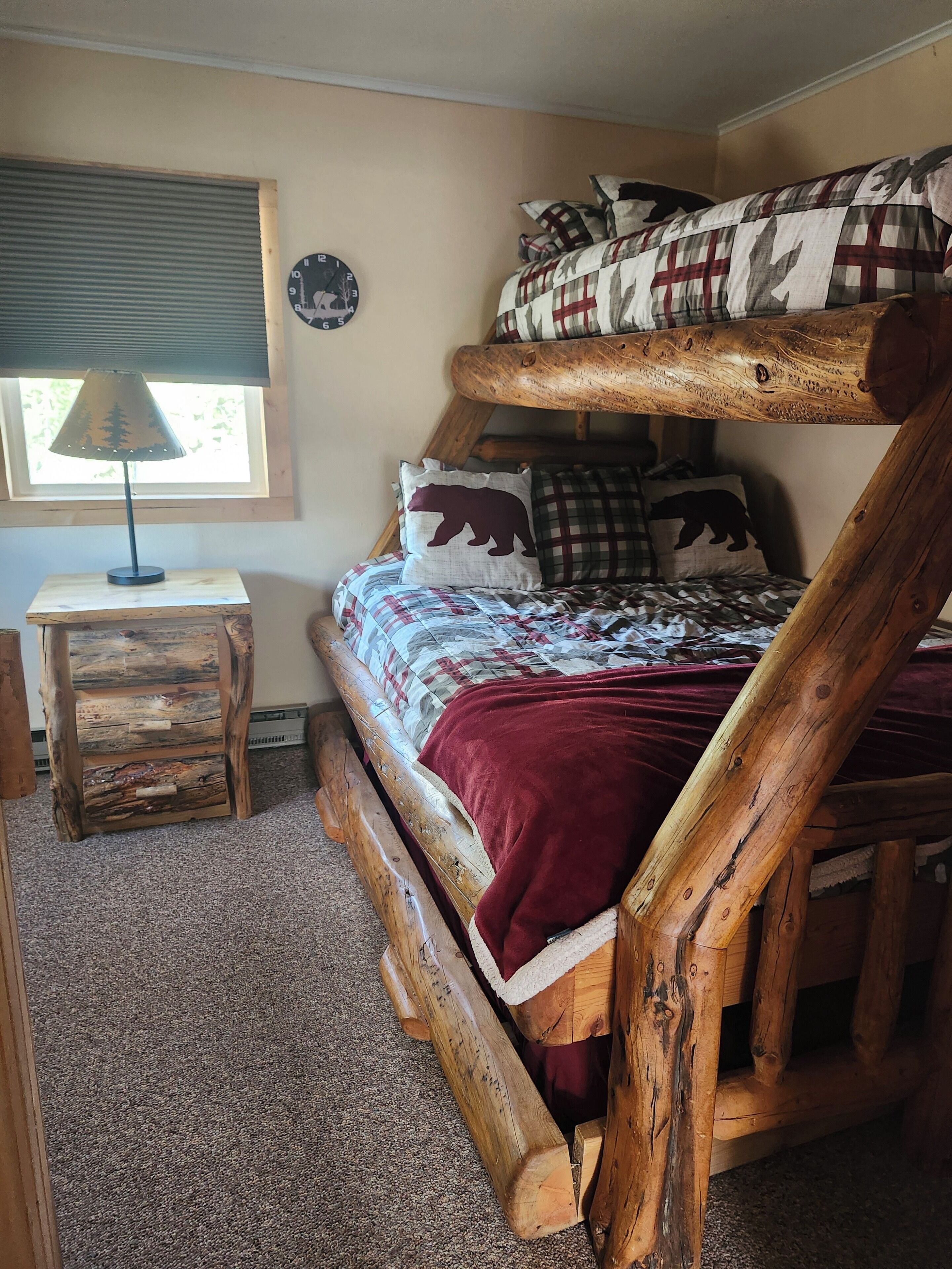 A bunk bed with a wooden frame, a bedside table with a lamp, and a window with blinds.