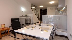 A modern dining area with a white table set for four, a black chair, a wooden side table, and a kitchen in the background. - Parque Santiago II 85 - Two Bed south facing (Playa de la Américas)