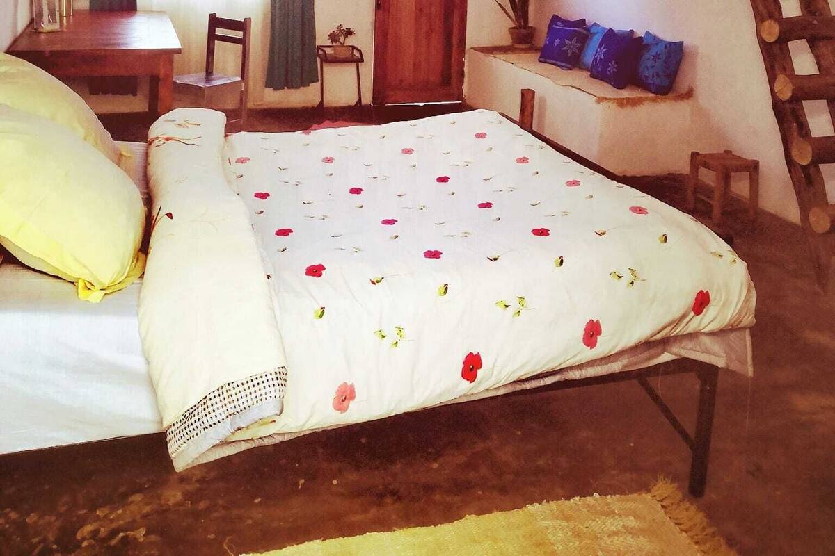 A rustic bedroom with a wooden bed, a small table, a chair, a window with curtains, and a wooden ladder.