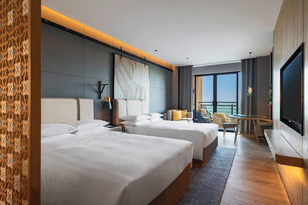 A hotel room with two beds, a large window, a TV, and a balcony with a view.
