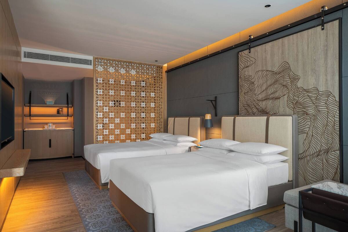 A modern hotel room with two beds, a seating area, a kitchenette, and decorative wall panels.