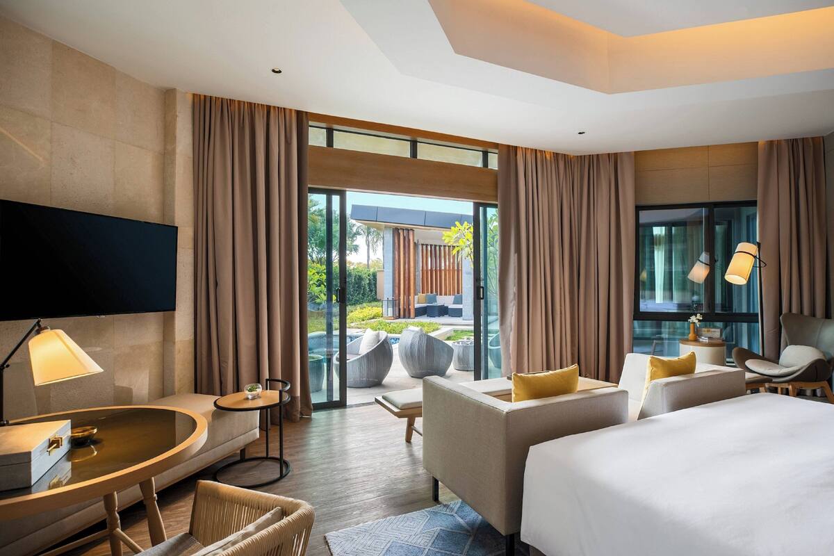 A modern hotel room with a large bed, a sofa, a desk, and a TV. There is a glass door leading to an outdoor area with plants and seating.