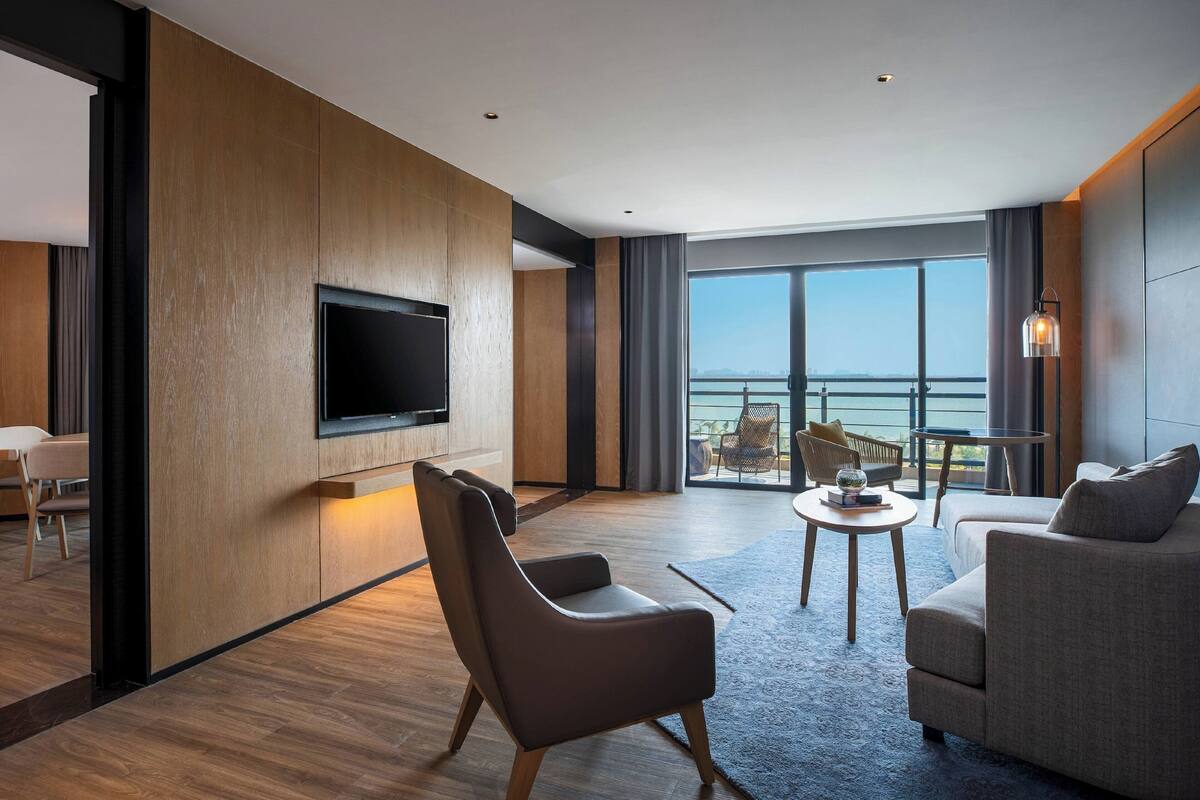 A modern hotel room with a large TV, comfortable seating, a small table, and a balcony with a view.