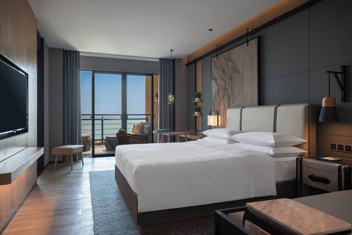 A modern hotel room with a large bed, a desk, a TV, a balcony with a view, and a large artwork on the wall.