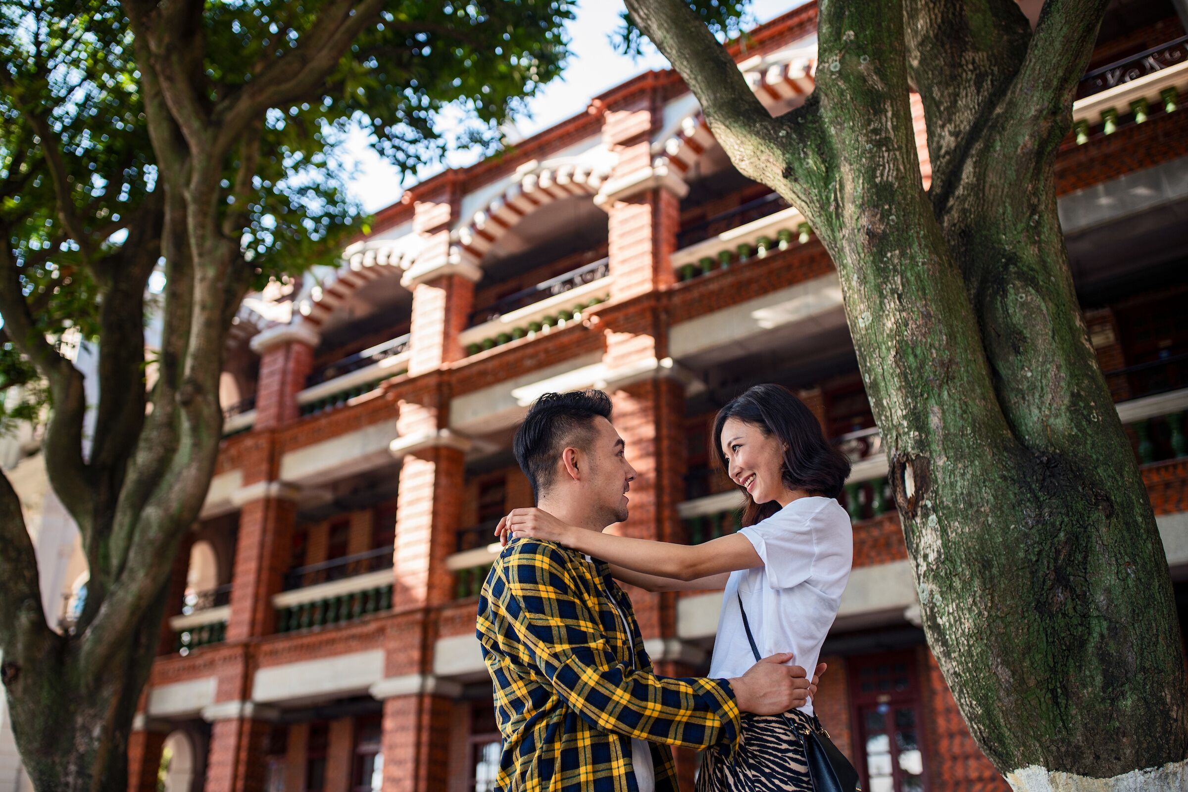 A couple embracing in front of a traditional building with multiple balconies and large trees.
