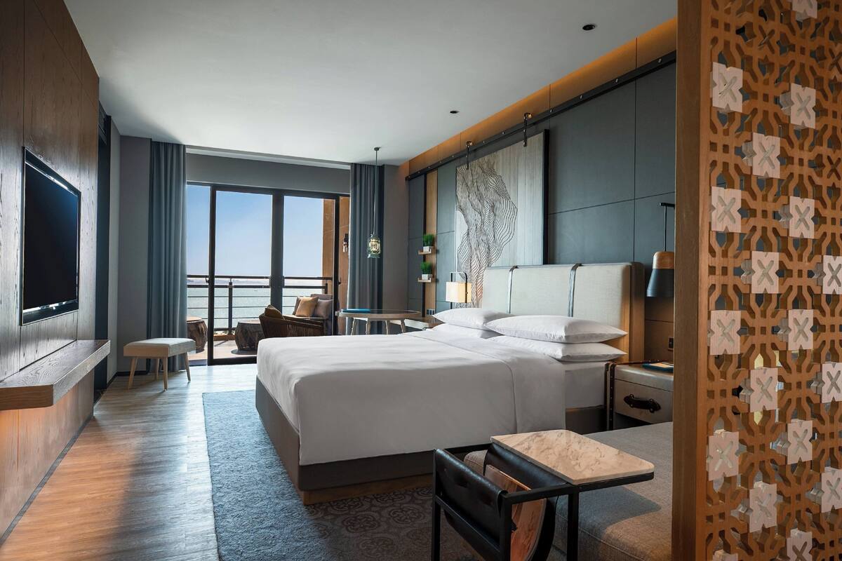 A modern hotel room with a large bed, a desk, a TV, and a balcony with a view.