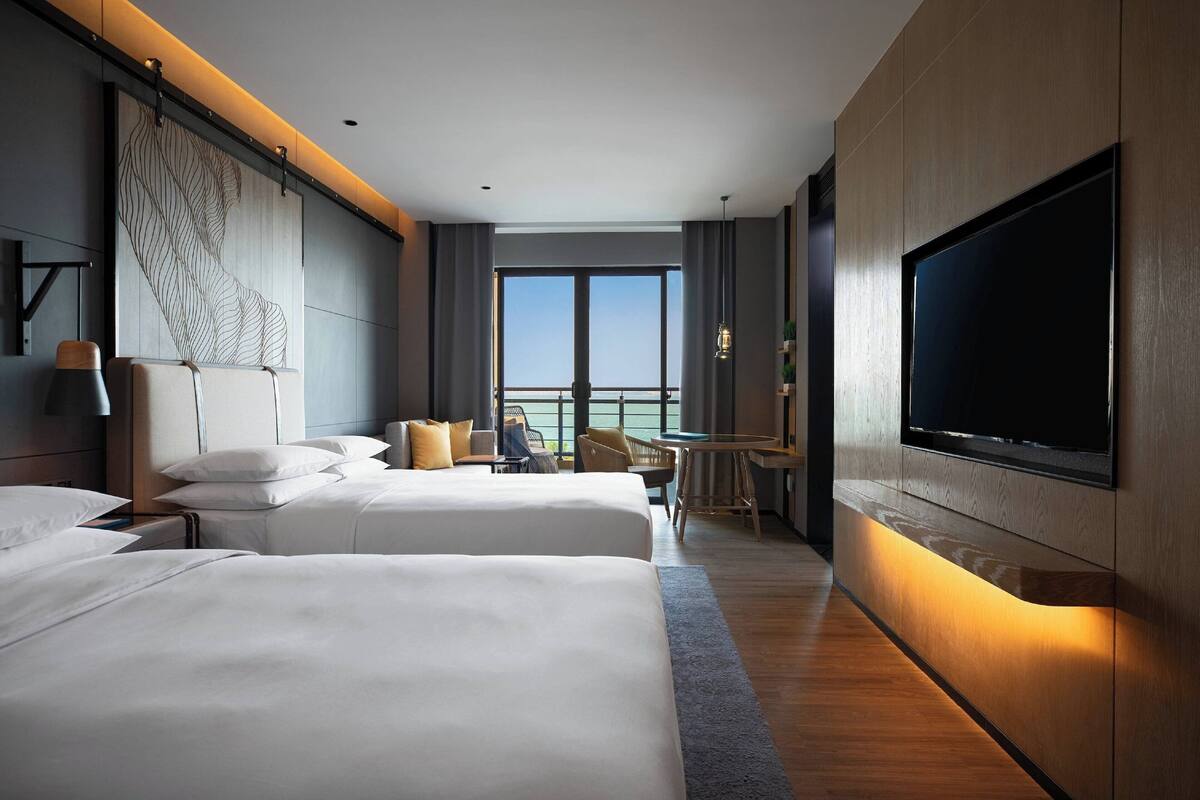 A hotel room with two beds, a large TV, a desk, and a view of the sea.