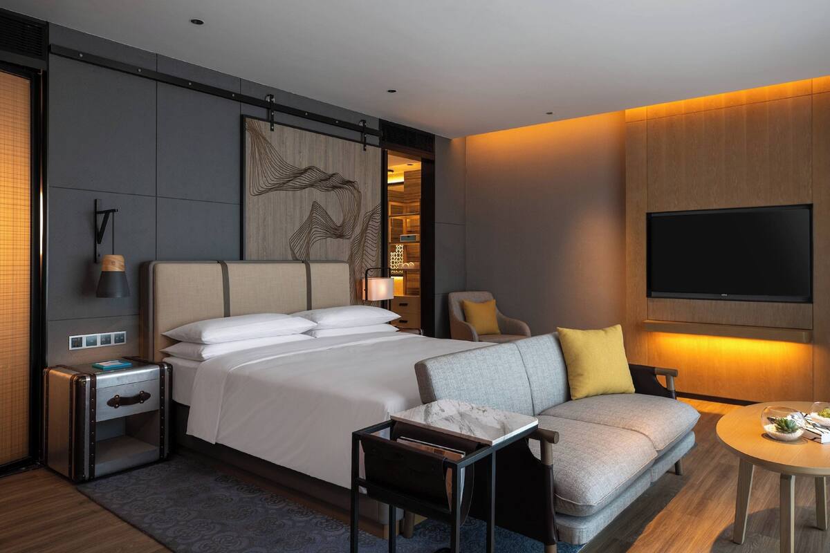 A modern hotel room with a large bed, a sofa, a small table, and a TV mounted on the wall.