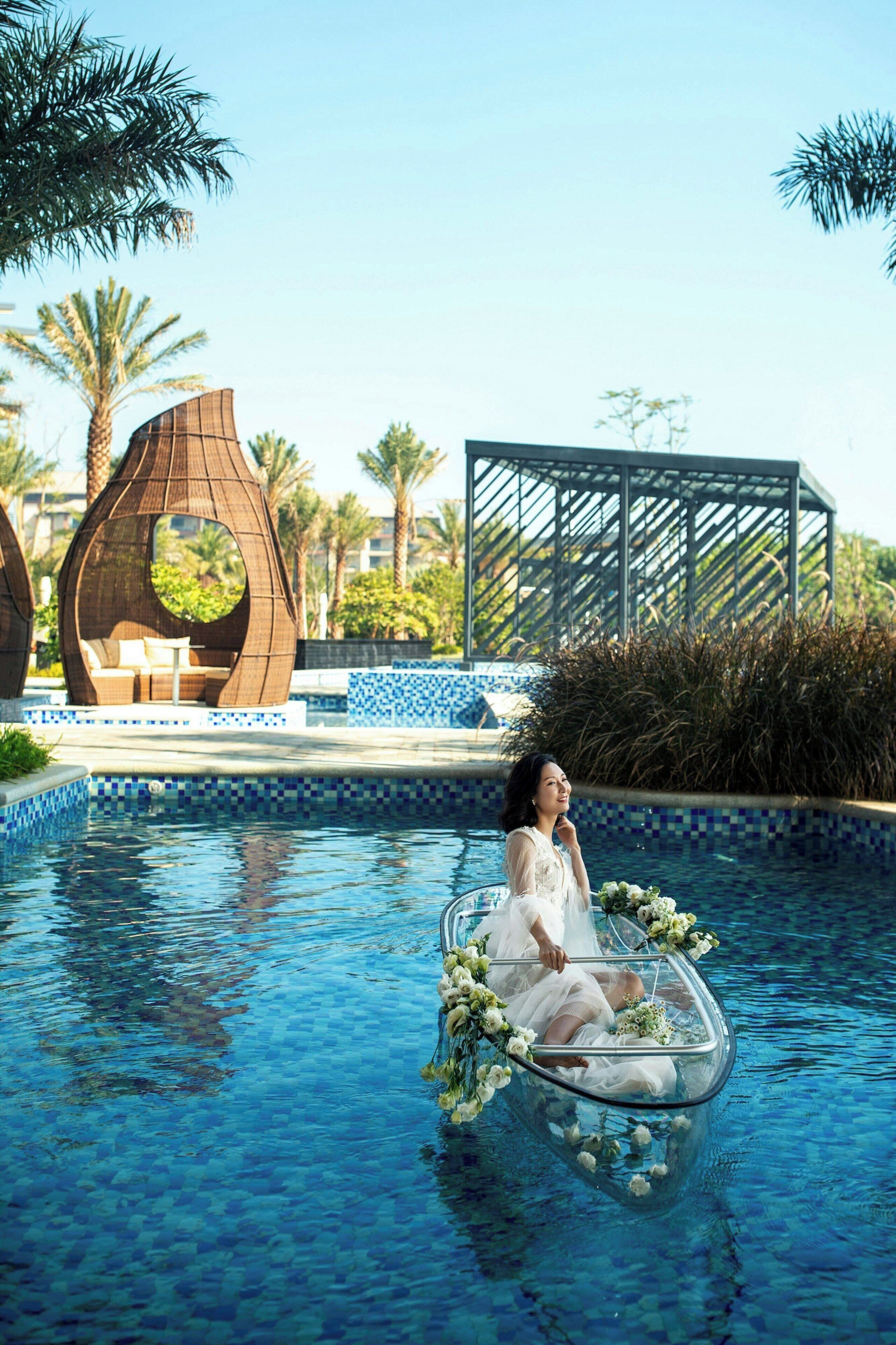 Une piscine avec un petit bateau en verre transparent, à l’intérieur duquel se trouve une personne vêtue d’une robe blanche, le tout entouré de fleurs.