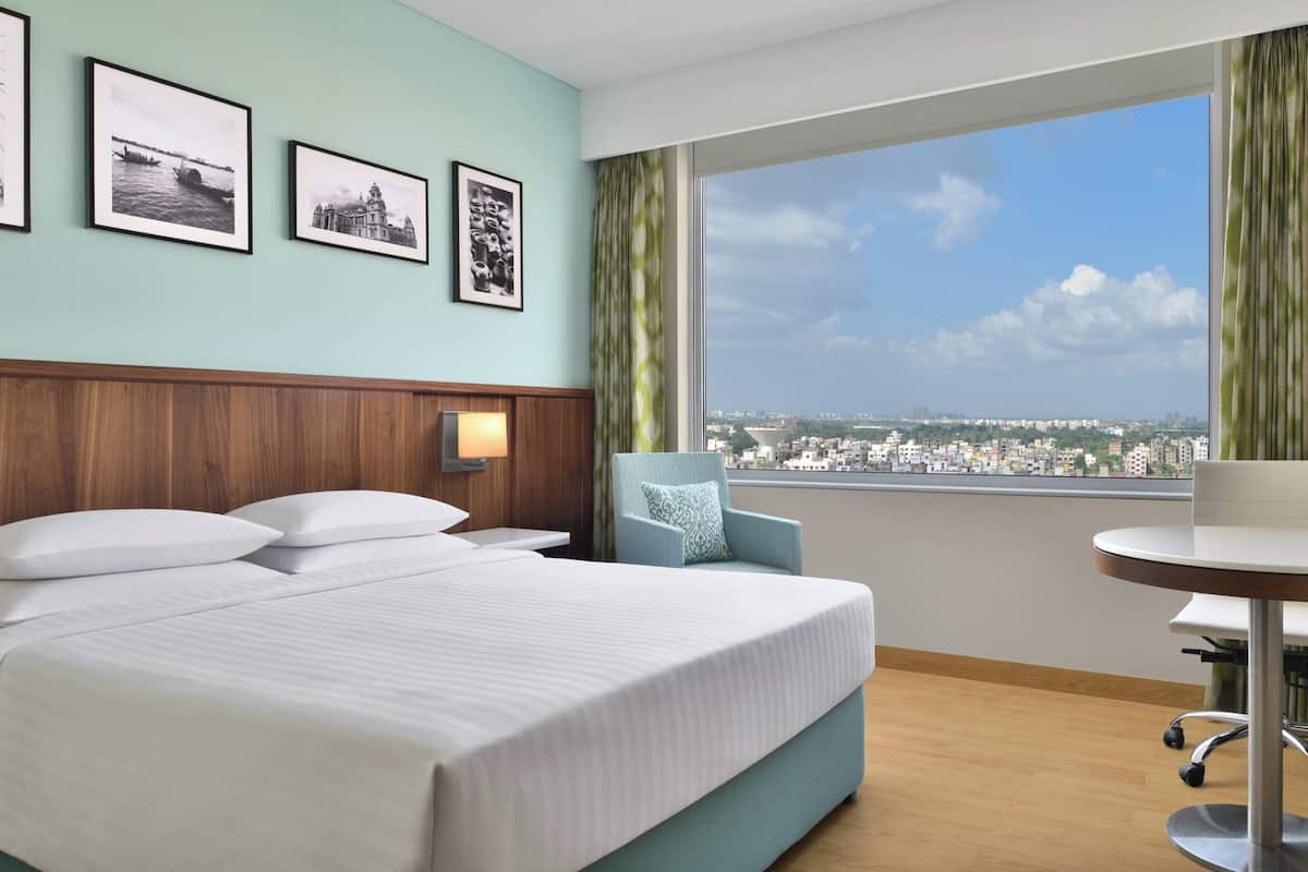 Habitación de hotel con cama, escritorio y vistas de un paisaje urbano a través de amplias ventanas.