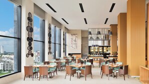 Un comedor moderno con varias mesas y sillas, ventanas amplias que ofrecen vistas a la ciudad, y una zona de bar con estantes llenos de botellas.