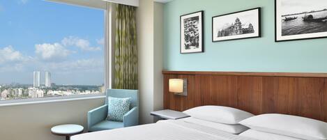 Habitación de hotel con una cama grande, una silla, una mesita y vistas a la ciudad a través de una ventana.