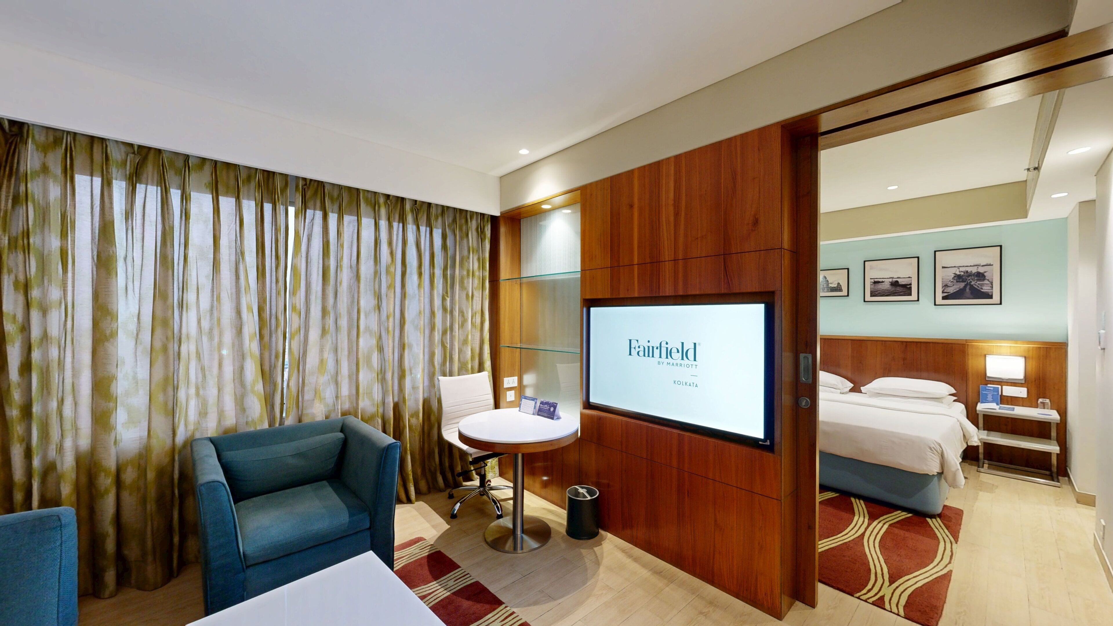 A hotel room with a bed, a desk, a chair, a TV screen displaying the hotel's name, and a view of the adjacent room.