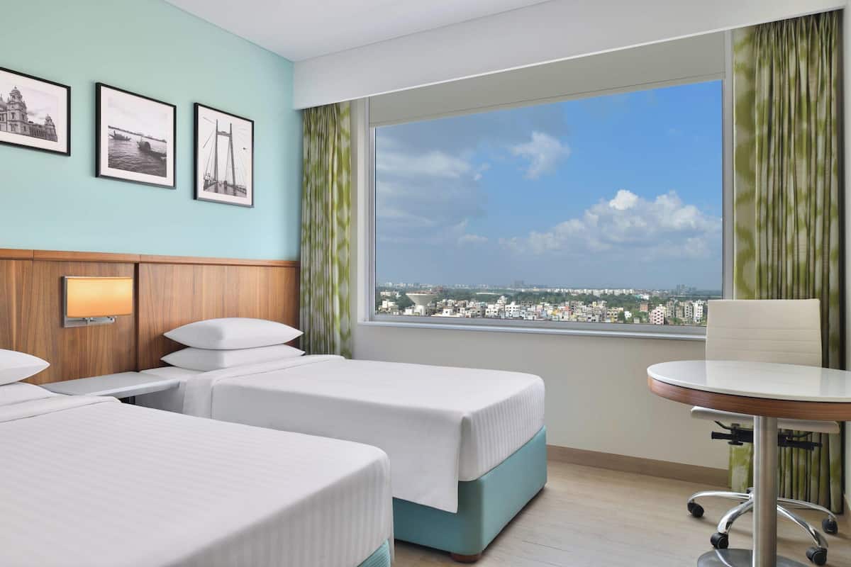 Habitación de hotel con dos camas, una mesita, una silla, un ventanal con vista a la ciudad y tres cuadros enmarcados en la pared.