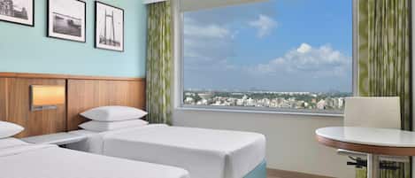 Ein Hotelzimmer mit zwei Betten, einem kleinen Tisch, einem Stuhl, einem Fenster mit Blick auf die Stadt und drei gerahmten Bildern an der Wand.