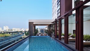 Una piscina en la azotea con vista a la ciudad, barandillas de vidrio y mobiliario exterior.