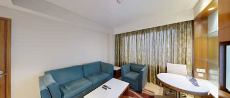 Quarto de hotel moderno com um sofá azul, uma mesinha branca, um aparador de vidro e uma cortina com padrão de folha.
