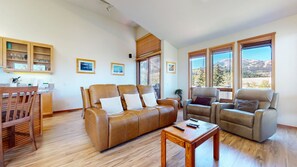 A spacious living room with a brown leather sofa, a wooden coffee table, and a grey armchair. There are framed pictures on the wall, a kitchen area with wooden cabinets, and large windows with a mountain view. - Snowcreek V 999 Mammoth Mountain Views, Private Washer Dryer, Pet-Friendly by RedAwning (Mammoth Lakes)