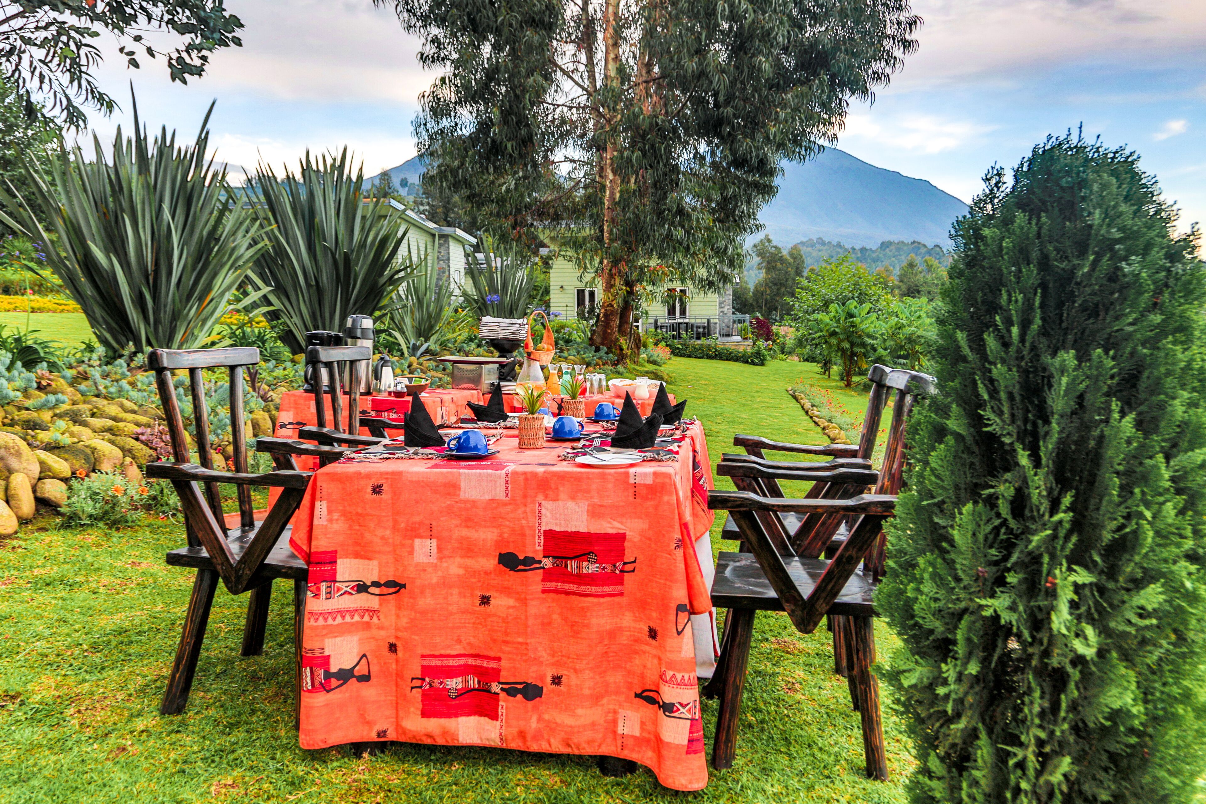 An outdoor dining area with a table set for four, surrounded by chairs, in a lush garden with a mountain in the background.