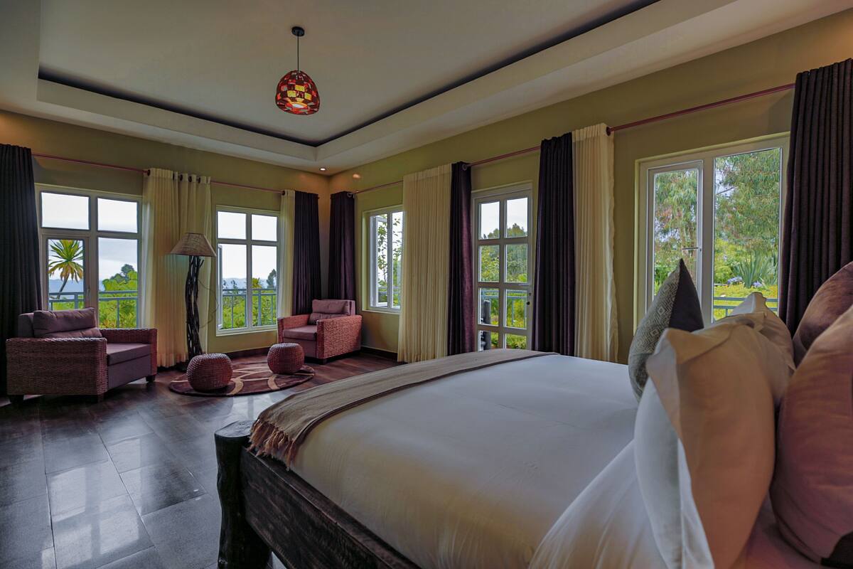 A spacious bedroom with a large bed, two armchairs, a floor lamp, and large windows offering a view of greenery.