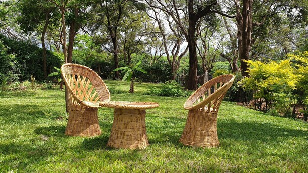 Uma área de estar externa em vime, com duas cadeiras e uma mesa, rodeada por vegetação exuberante e árvores.