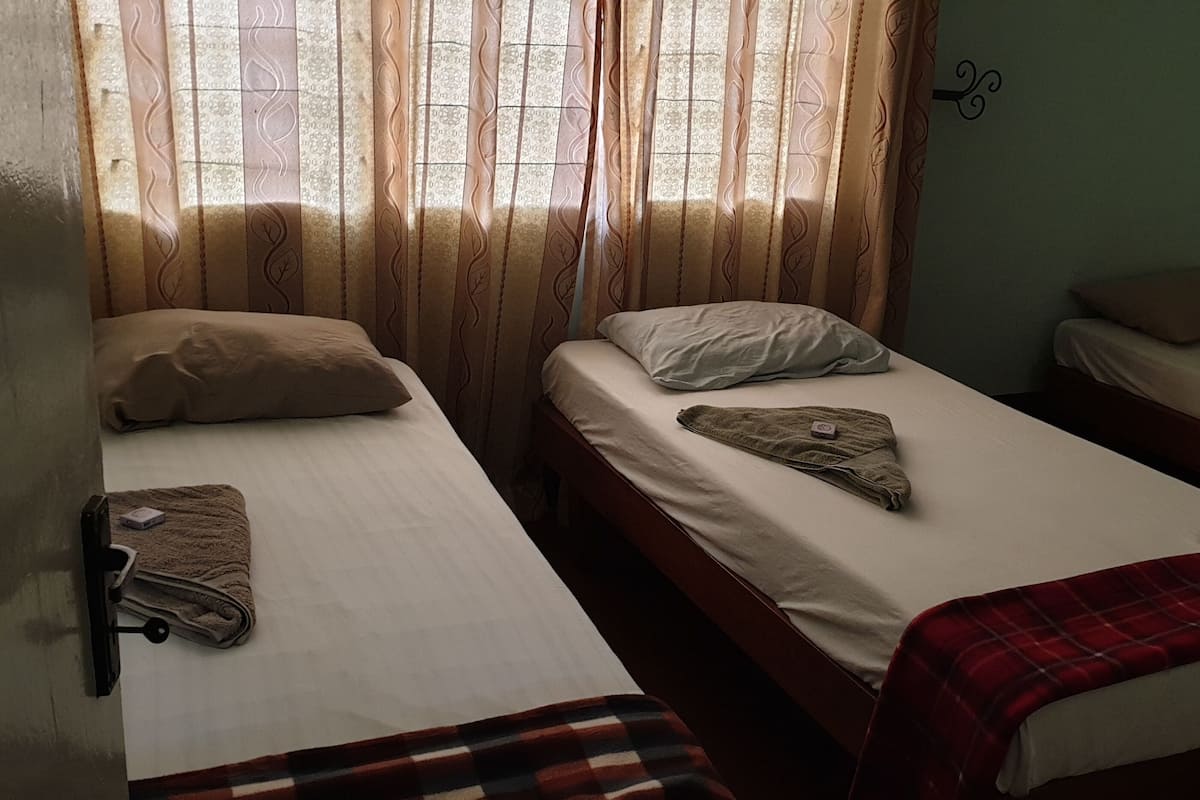 A room with two single beds, each with a pillow and a blanket. There is a window with curtains, and a wall-mounted light fixture.